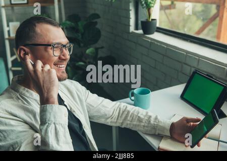 Kaukasischer Mann, der in seinem Büro mit seinem Telefon, Tablet und seinem Programm arbeitet. Moderne Kommunikation. Drahtlose Kommunikation. Moderner Lifestyle. Stockfoto