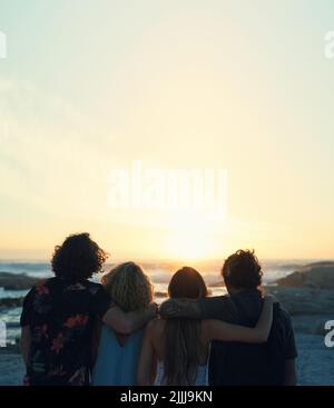Eine Gruppe von Freunden umarmen sich am Strand mit Blick auf den wunderschönen Sonnenuntergang und genießen den Sommerurlaub Stockfoto