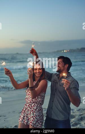 Glückliches Paar, das am Abend am Strand bei Sonnenuntergang mit funkelnden Feuerwerken die Neujahrsfeier zelebriert Stockfoto