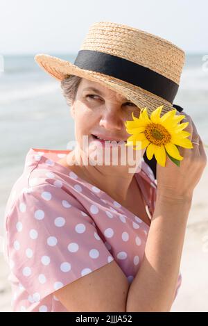 Glücklich verheiratete grauhaarige Frau in einem Bootsfahrer mit einer leuchtend gelben Blume, in einem rosa Kleid mit weißen Tupfen, die etwas mit Interesse beobachten. Stockfoto