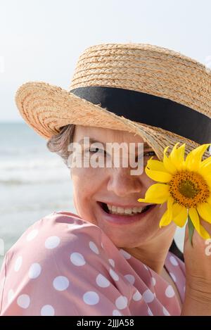 Fröhlich lächelnde grauhaarige Frau in einem Bootsfahrer und rosa Kleid mit weißen Punkten mit einer gelben Blume, die auf die Kamera schaut, Nahaufnahme Porträt.