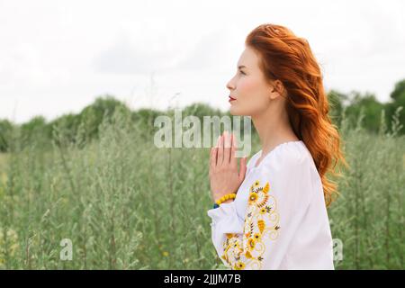 Die rothaarige, schöne, junge Frau in einem ukrainischen bestickten Hemd betet für die Ukraine auf dem Feld Stockfoto