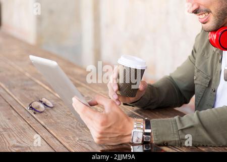 Zugeschnittenes Foto eines erwachsenen Mannes, der Kaffee trinkt, während er ein digitales Tablet auf einer Terrasse im Freien verwendet Stockfoto