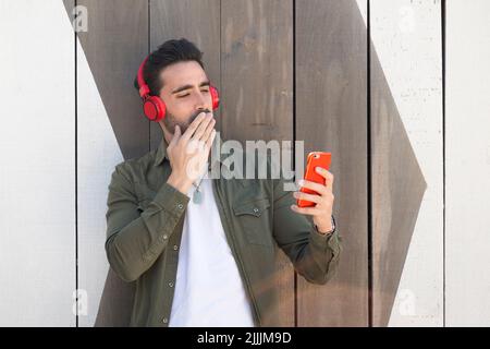 Überraschte den Mann, der das Handy benutzte, während er Musik auf der Straße neben einer Holzwand hörte Stockfoto