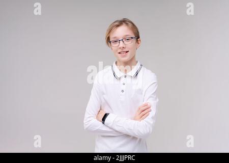 Zuckender kaukasischer Junge, zweifelende zuckende Schultern, Schuljunge, Teenager mit Brille in einem weißen T-Shirt. Der Junge wählt auf einem grauen Hintergrund. Stockfoto