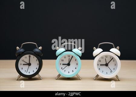 Drei kleine Wecker schwarz blau und weiß zeigen neun Uhr, stehen auf einem Tisch auf schwarzem Hintergrund Stockfoto