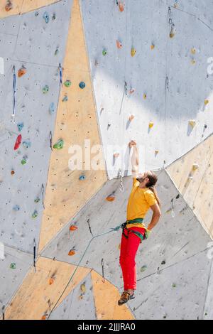 Sportkletterer hängt an einer Hand an einer künstlichen Kletterwand im Freien. Der Mann schob seine Hände hinter seinem Rücken in einen Beutel mit Magnesiuumpulver. Stockfoto