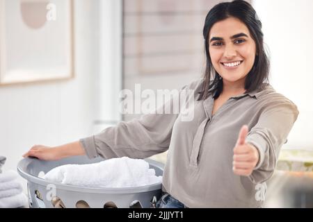 Eine junge Frau zeigt den Daumen nach oben, während sie zu Hause Wäsche macht. Stockfoto