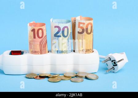 Euro-Scheine in Steckerleiste und Stecker mit Münzen auf blauem Hintergrund. Konzept der teuren Stromkosten in Europa und der steigenden Energiebillimpreise. Stockfoto