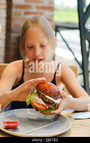 Fröhliches Mädchen, Teenager, die in einem Restaurant auf einer Sommerterrasse einen Burger mit Fleischpastete essen. Essen und Lifestyle. Fast Food. Mittagessen Stockfoto