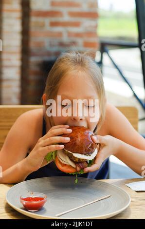 Fröhliches Mädchen, Teenager, die in einem Restaurant auf einer Sommerterrasse einen Burger mit Fleischpastete essen. Essen und Lifestyle. Fast Food. Mittagessen Stockfoto