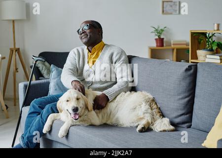 Älterer blinder Mann, der während seiner Freizeit auf dem Sofa zu Hause mit dem Blindenhund spielt Stockfoto