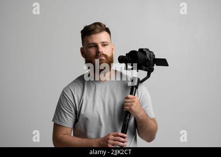 Gutaussehender bärtiger Videofilmer, der mit Steadicam ein gutes Filmmaterial macht. Der Kameramann, der einen Stabilisator in der Hand hat, nimmt ein Video auf dem slr-Video auf Stockfoto