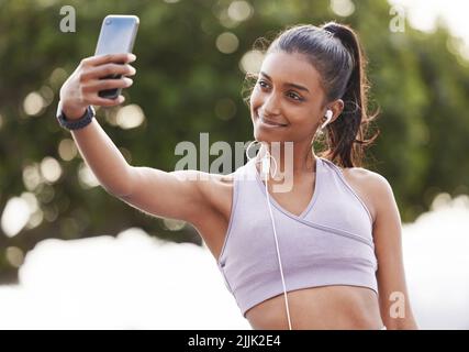 Ich bekomme meinen Körper in die beste Form. Eine sportliche junge Frau, die Kopfhörer trägt und Selfies macht, während sie draußen trainiert. Stockfoto