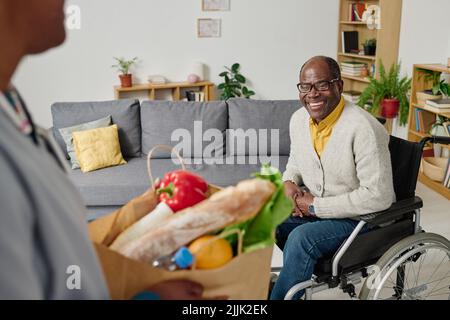 Afrikanischer glücklicher Mann mit Behinderung, der im Rollstuhl sitzt und einen Betreuer trifft, der Lebensmittel für ihn bringt Stockfoto