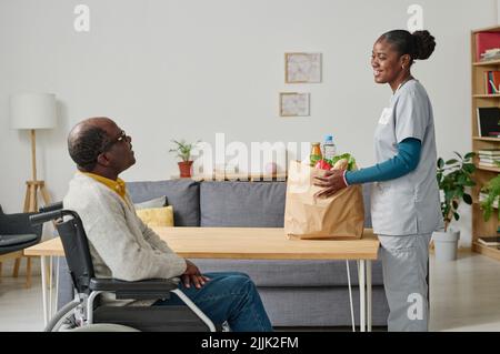 Afrikanische junge Freiwillige bringt Essen in Papiertüte für ältere Mann im Rollstuhl, sie lächelt und spricht mit ihm im Zimmer Stockfoto