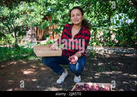 Die erfolgreiche Amateurfarmerin blickt lächelnd auf die Kamera, während sie geerntete Kartoffeln im Hinterhof ihres Landhauses sortiert Stockfoto