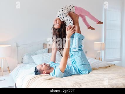 Wache auf und erlebe einen vergnüglichen Tag. Ein Mann, der auf einem Bett liegt und seine Tochter mit den Füßen in die Luft hebt. Stockfoto