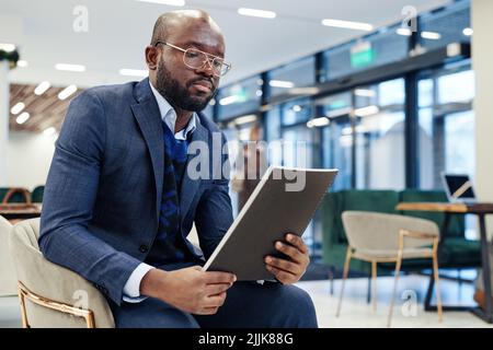 Afrikanischer Geschäftsmann in formellem Anzug, der sich auf das Lesen des Berichts konzentriert, in der Lobby sitzt und sich auf das Treffen vorbereitet Stockfoto