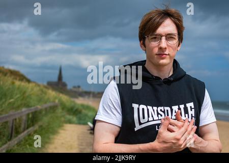 Natürliches Halbportrait eines jungen Mannes mit ärmellosem Hoodie und weißem T-Shirt am Strand in Tynemouth mit umschlingenden Händen Stockfoto