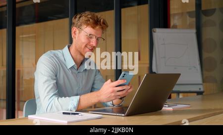 Lächelnder junger erwachsener europäischer Geschäftsmann, der im Büro ein Mobiltelefon benutzt. Stockfoto