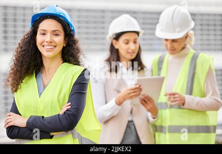 Das Management ist am Job. Zugeschnittenes Porträt einer attraktiven jungen Ingenieurin, die mit gefalteten Armen und ihren Kollegen im Hintergrund steht Stockfoto