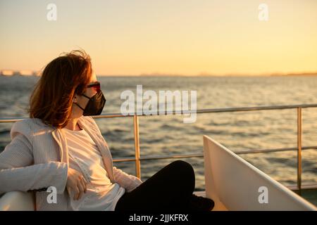 Eine junge Frau mit medizinischer Gesichtsmaske und Sonnenbrille auf einem Pendlerboot bei Sonnenuntergang Stockfoto