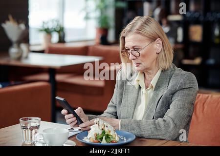 Reife Geschäftsfrau im Anzug mit Mobiltelefon beim Mittagessen am Tisch im Café Stockfoto