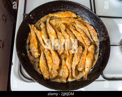 Draufsicht auf das Braten kleiner Schwarzer Meerbarsche in alter, runder gusseiserner Bratpfanne auf einem Gasherd in der rustikalen Küche Stockfoto