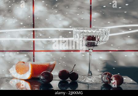 Kristallglas für Cocktails auf einer Glasoberfläche. Früchte - Trauben, Grapefruit neben dem Glas. Abstrakter Hintergrund. Stockfoto