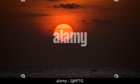 Großer Ball der orangefarbenen Sonne untergeht hinter dem Ozeanhorizont, wunderschöne Sonnenuntergangsfarben am Himmel. Einsame Silhouette Fischerboot segelt im Meer. Stockfoto