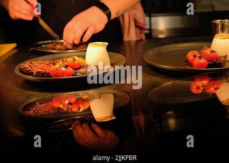 Köche bereiten in einer kommerziellen Küche Teller mit Lebensmitteln für die Kunden zu. Rindersteak, Weintomaten, Pilze, Pfeffersauce auf dem Teller. Stockfoto