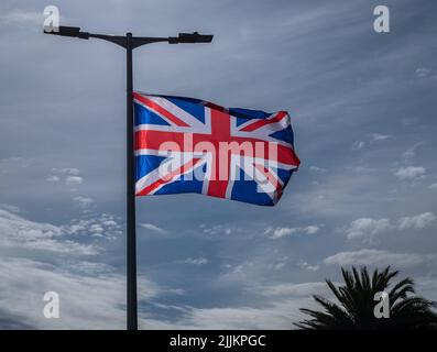 Britische Flagge auf Fahnenmast gegen blauen Himmel mit heller Wolke. Flagge im Wind, von der Sonne beleuchtet. Leuchtend rote und blaue Farben mit Palmen. Stockfoto