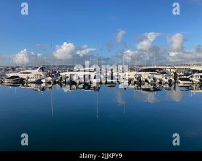 Segelboote und Motoryachten an einem sonnigen Tag in einer Marina. Boote und Masten spiegelten sich im stillen Wasser in einer ruhigen Szene wider. Flauschige Wolken am Himmel. Stockfoto