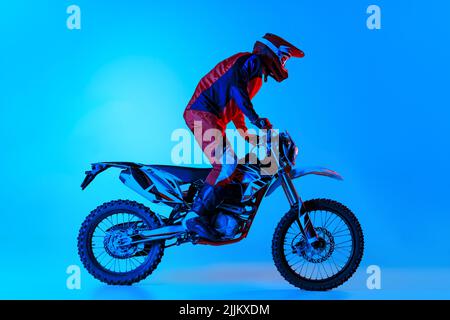 Porträt eines jungen Mannes, Biker, der auf einem Motorrad steht, isoliert über blauem Studiohintergrund in Neonlicht reitet Stockfoto