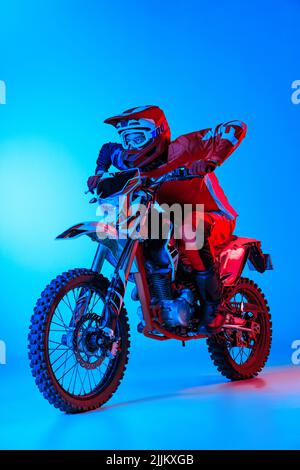 Porträt eines jungen Mannes, der auf einem Motorrad auf blauem Studiohintergrund in Neonlicht reitet. Sporttraining Stockfoto