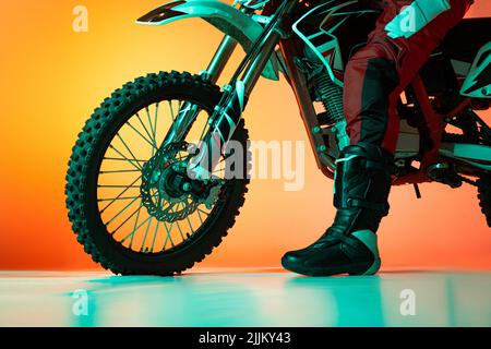 Zugeschnittenes Bild von einem Mann, einem Biker-Bein in Stiefeln und einem Motorrad-Reifen, isoliert über einem orangen Studiohintergrund in Neonlicht Stockfoto