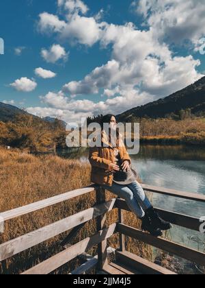 Eine junge Frau, die im Herbst auf einem Holzdeck am idyllischen See im Naturschutzgebiet Zelenci in Slowenien sitzt Stockfoto