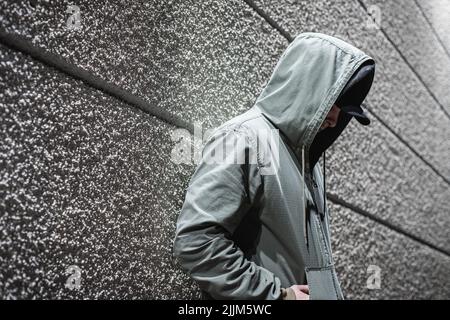 Eine Seitenansicht einer Person in einem Anorak mit den Händen in den Taschen, die an einer gestreiften Wand gelehnt sind Stockfoto