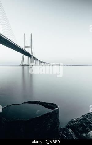 Eine vertikale Graustufenaufnahme der Vasco da Gama Brücke mit Spiegelung im Wasser und grauem Himmel Stockfoto