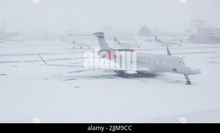 Schneebedecktes Verkehrsflugzeug am Flughafen. Nahaufnahme des Flugzeugs nach Schneefall. Flughafenmitarbeiter reinigen Start- und Landebahnen. Winter schlechte Wetterbedingungen. Stockfoto