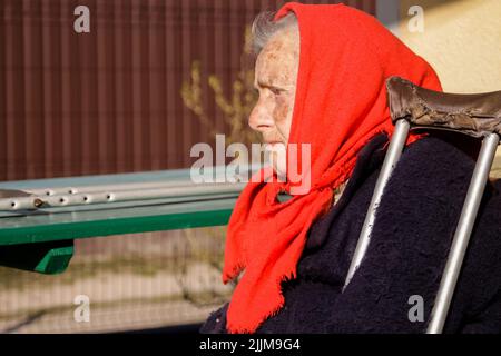 Defokussieren sie die ältere ukrainische Frau in einem roten Schal mit Krücken, die draußen sitzen. Frau 85 Jahre alt. Einzelperson. Nahaufnahme Porträt. Krank und heimatverbunden. Cop Stockfoto