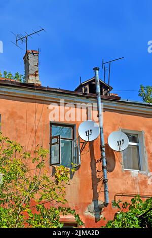 Wenige Fernseher und Multi-Feed-Satellitenantenne mit mehreren Feeds (Konverter) an der alten roten Gebäudewand mit offenen Fenstern und hohem silbernen ME Stockfoto