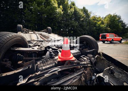 Beschädigtes Fahrzeug auf dem Dach nach einem Unfall mit einem Feuerwehrfahrzeug. Selektiver Fokus auf den Verkehrskegel. Stockfoto