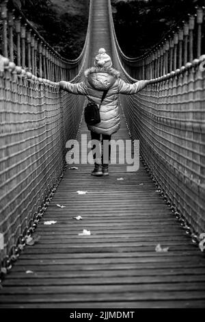 Eine vertikale Graustufenaufnahme einer Person, die auf einer Hängebrücke, umgeben von Bäumen in einem Wald, läuft Stockfoto