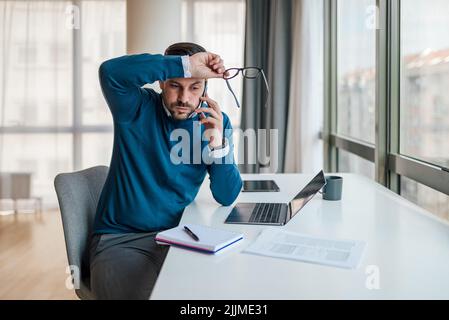 Besorgt professionelle Gespräche auf dem Handy. Männlicher Unternehmer arbeitet am Schreibtisch am Laptop. Er sitzt am Fenster im Büro. Stockfoto