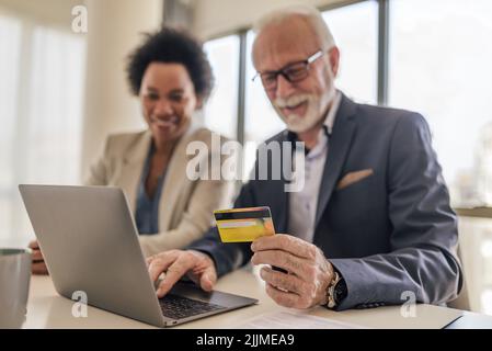 Nahaufnahme eines Geschäftsmannes, der online über einen Laptop bezahlt. Ältere männliche Berufstätige arbeiten mit weiblichen Kollegen zusammen. Der Schwerpunkt liegt auf der Kreditkarte Stockfoto