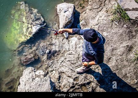 2022 06-25 Anchorage Alaska USA - Ansicht von oben nach unten von einem nicht erkennbaren Mann mit Mütze, der in Ship Creek in der Nähe der Innenstadt nach Lachs fischt. Stockfoto