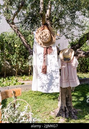 Frau und Kind Sommerkleider und Strohhüte hingen unter Olivenbaum, Stuhl im Garten, Korb mit Oliven-Brunches. Stockfoto