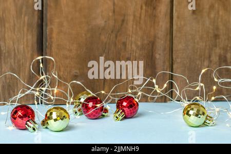 Ein Schuss goldener und roter Weihnachtskugeln mit einer Lichterkette auf einem weißen Tisch an der braunen Holzwand Stockfoto